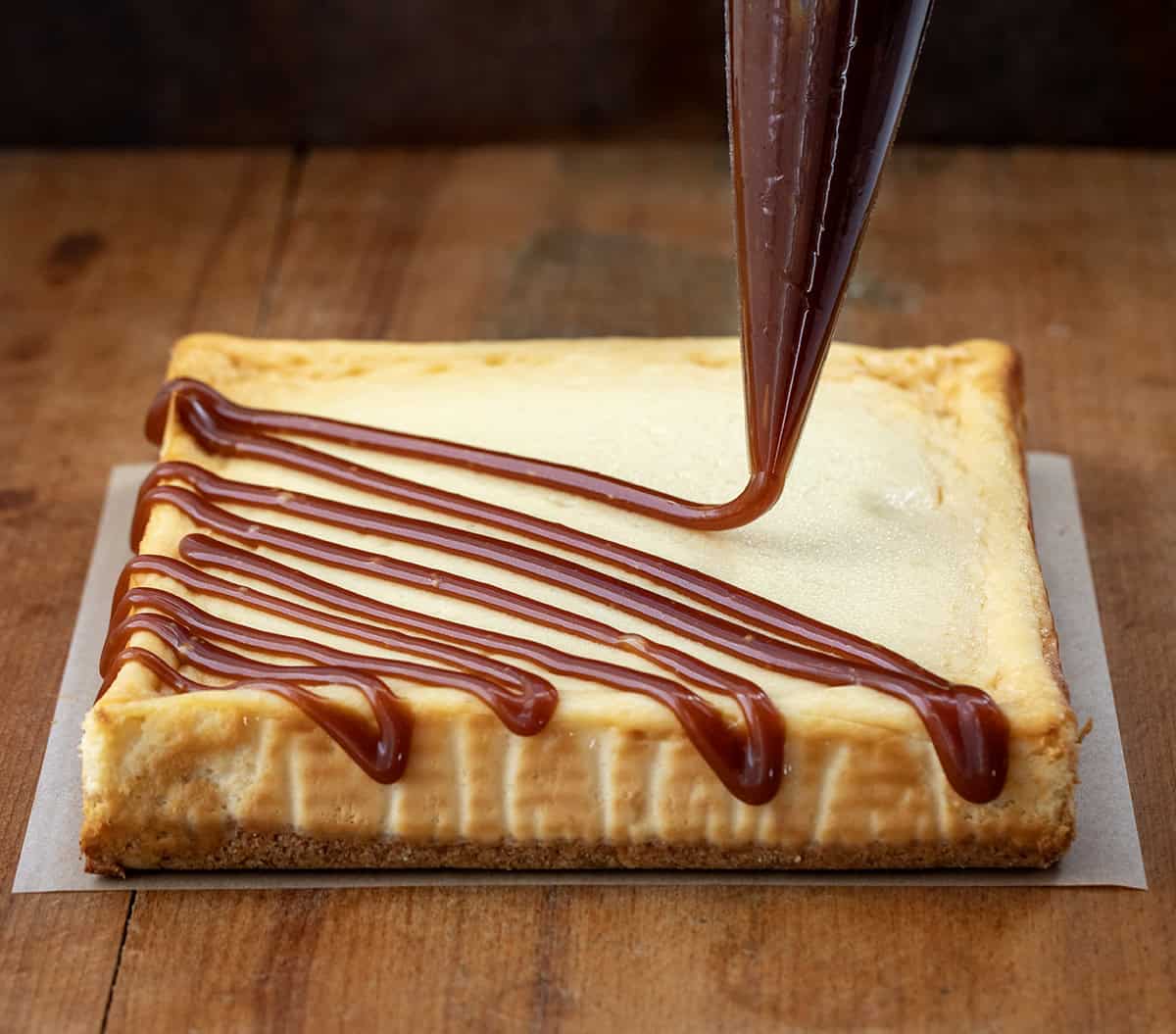Drizzling apple cider caramel over apple cider cheesecake bars on a wooden table.