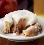 Portion of an Apple Cider Cinnamon Roll on a white plate with apple cider and apples in the background.