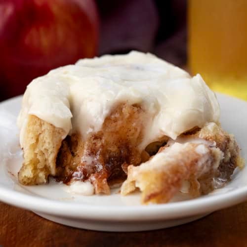 Portion of an Apple Cider Cinnamon Roll on a white plate with apple cider and apples in the background.
