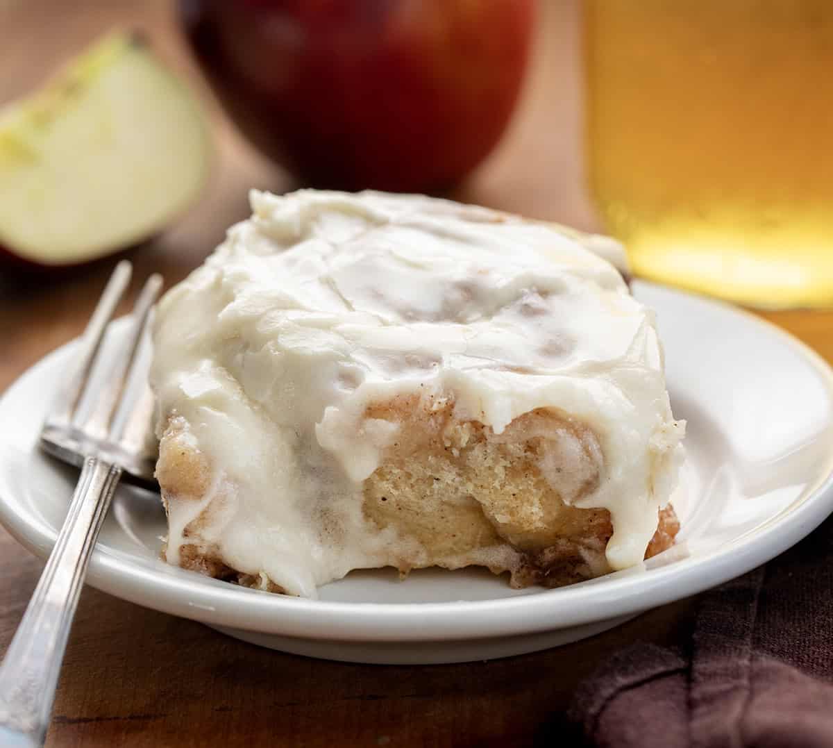 Apple Cider Cinnamon Roll on a white plate with apple cider and apples in the background.