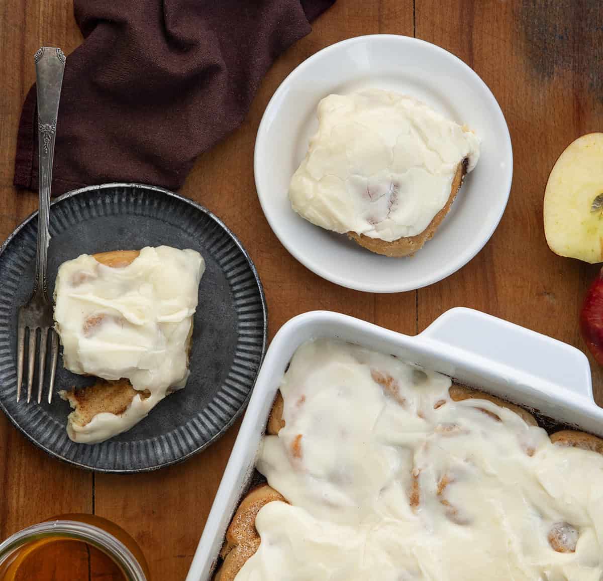 Apple Cider Cinnamon Rolls in a pan with a couple of rolls on plates near the pan.