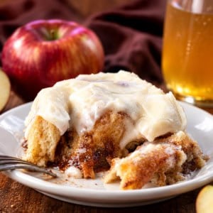 Portion of an Apple Cider Cinnamon Roll on a white plate with apple cider and apples in the background.