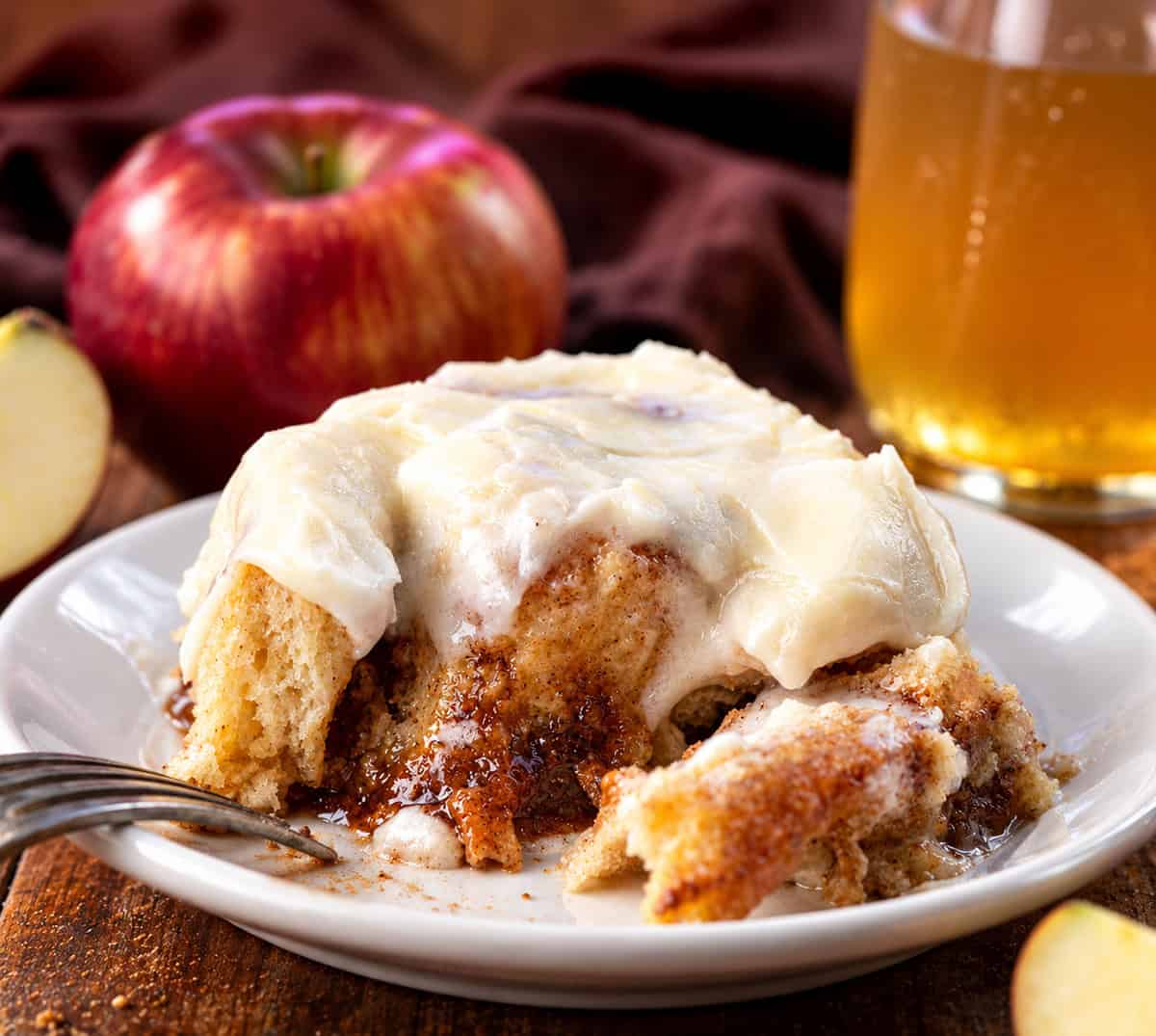 Portion of an Apple Cider Cinnamon Roll on a white plate with apple cider and apples in the background.
