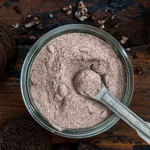 Looking down on a jar of Cookies & Cream Hot Cocoa Mix with a measuring spoon in it on a wooden table from overhead.