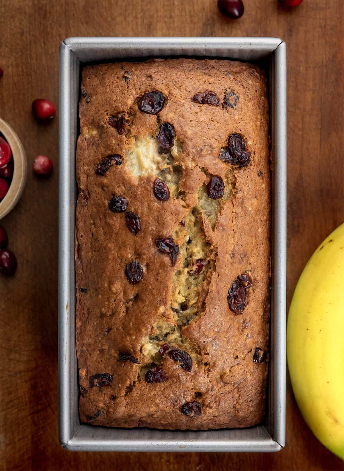 Loaf of Cranberry Banana Bread on a wooden table with fresh cranberries and bananas from overhead.