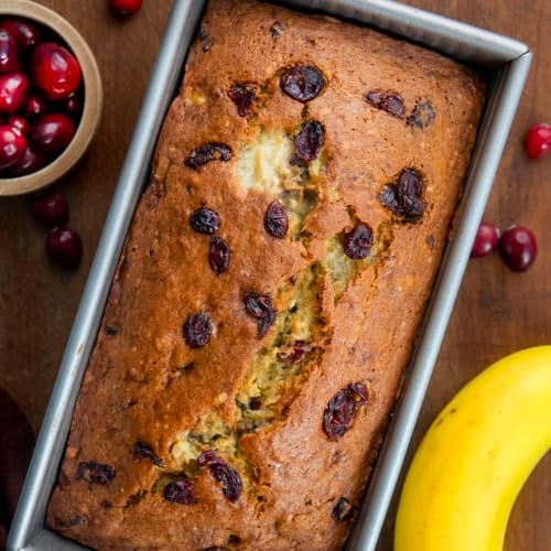 Loaf of Cranberry Banana Bread on a wooden table with fresh cranberries and bananas from overhead.